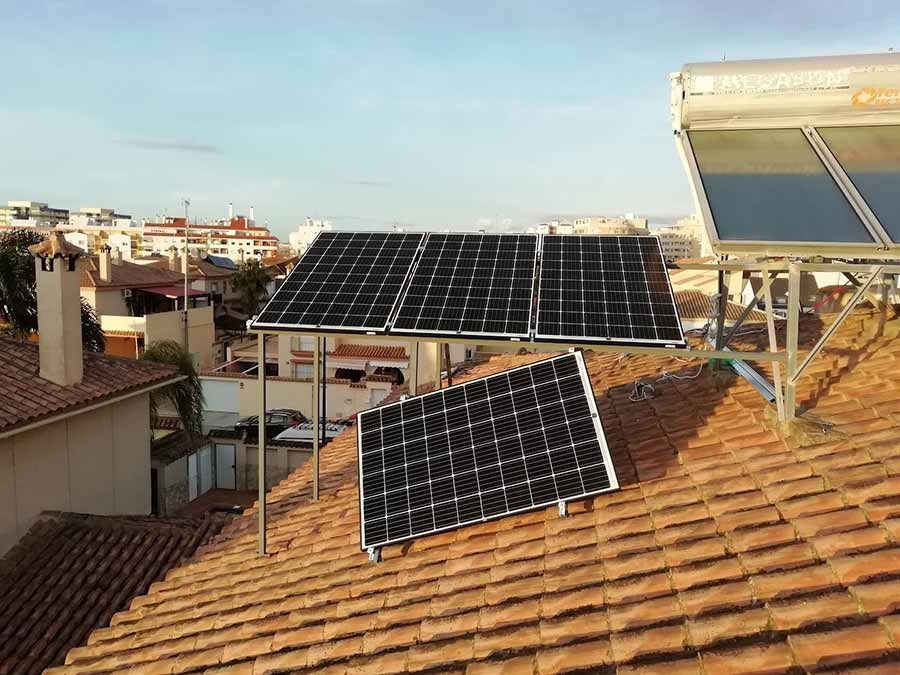 Paneles solares sobre un techo de tejas rojas con un calentador de agua, con vistas a un paisaje urbano bajo un cielo azul.