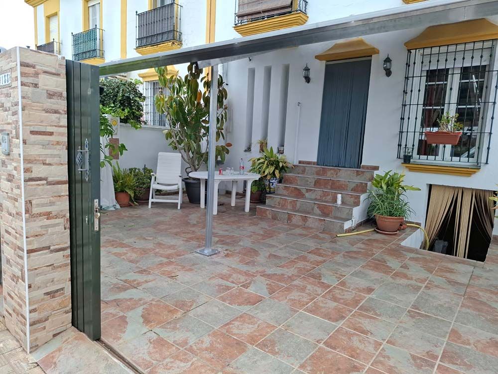 Patio con suelo de baldosas, portón, mesa, sillas, plantas y edificio con escalones que conducen a la entrada.