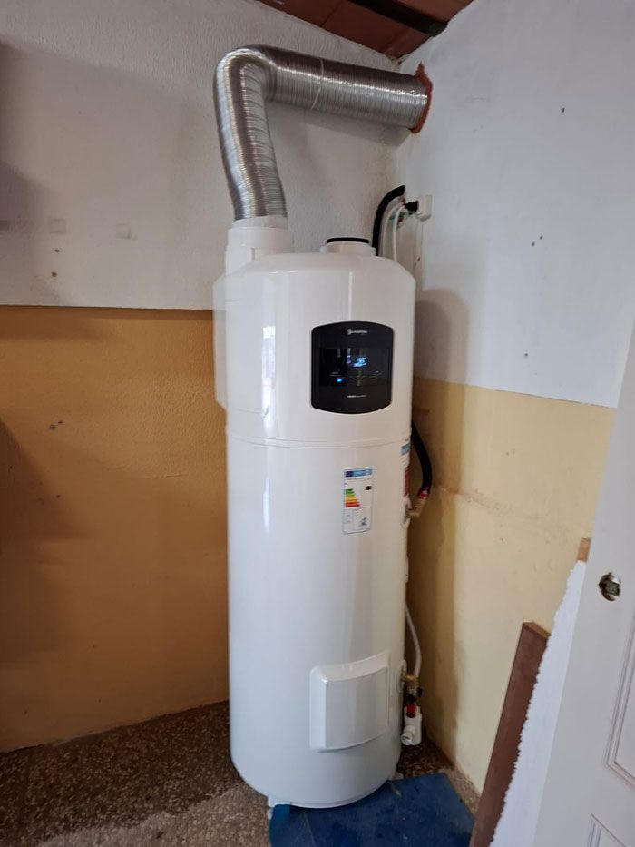 Calentador de agua blanco con tubo de ventilación plateado en una esquina de una habitación con paredes amarillas y blancas.