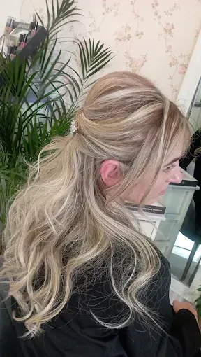 Mujer rubia de cabello ondulado vista de espaldas, con una blusa negra, en el interior de una casa cerca de unas plantas.