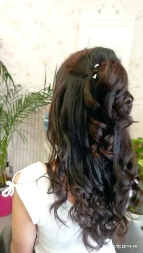 Mujer vista de espaldas con cabello largo, oscuro y ondulado, y una pinza blanca para el cabello, en el interior de una casa cerca de una planta.