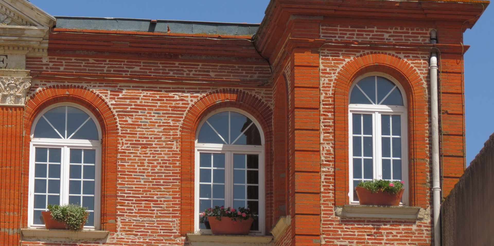 Bâtiment en briques rouges avec des fenêtres cintrées, des encadrements blancs et des jardinières sous un ciel bleu.