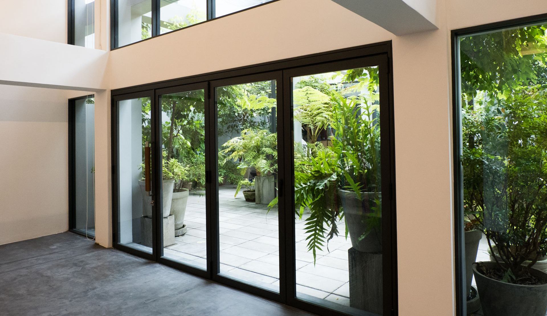 De grandes portes vitrées à cadre noir s'ouvrent sur une cour intérieure verdoyante, aux murs blancs et au sol gris.