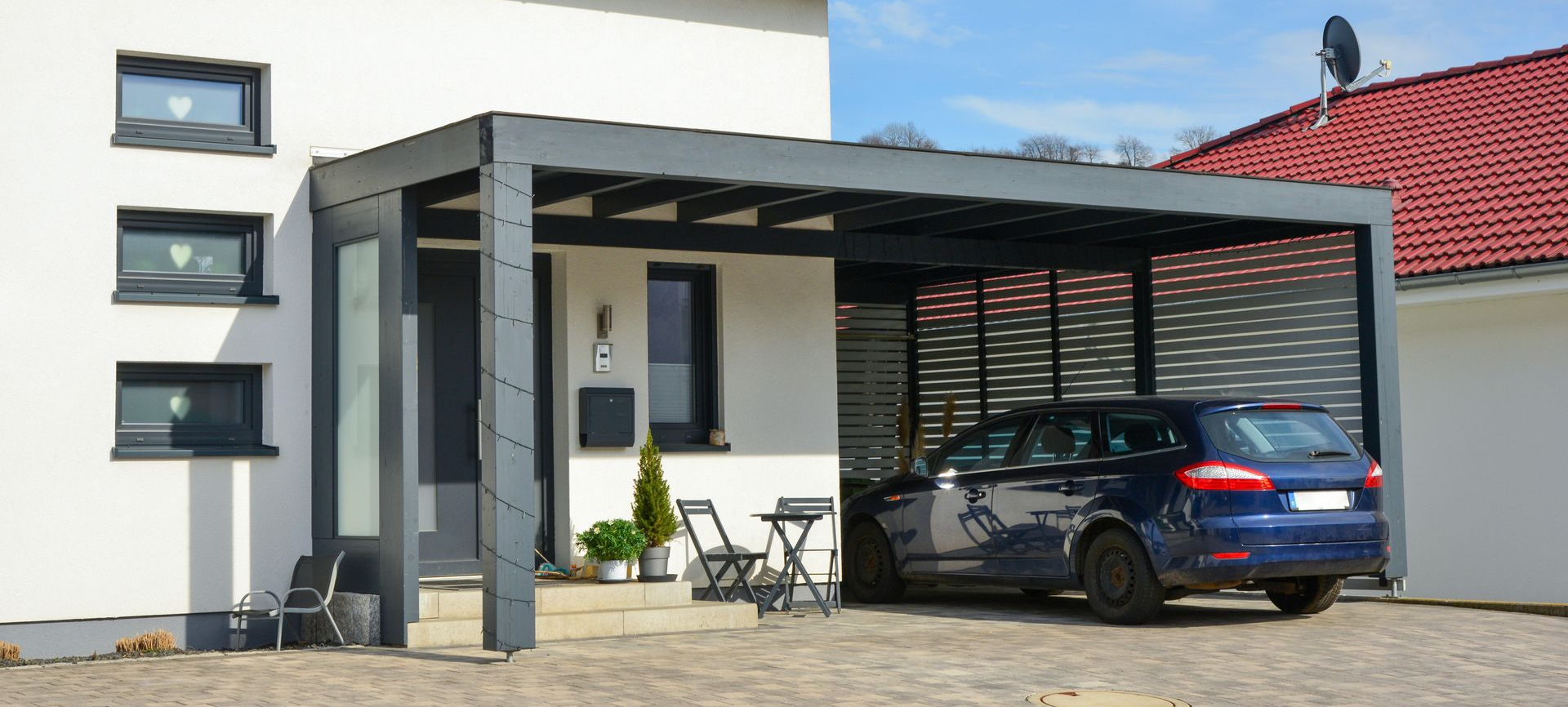 Un abri de voiture moderne est attenant à une maison blanche, avec une voiture bleue garée à l'intérieur.