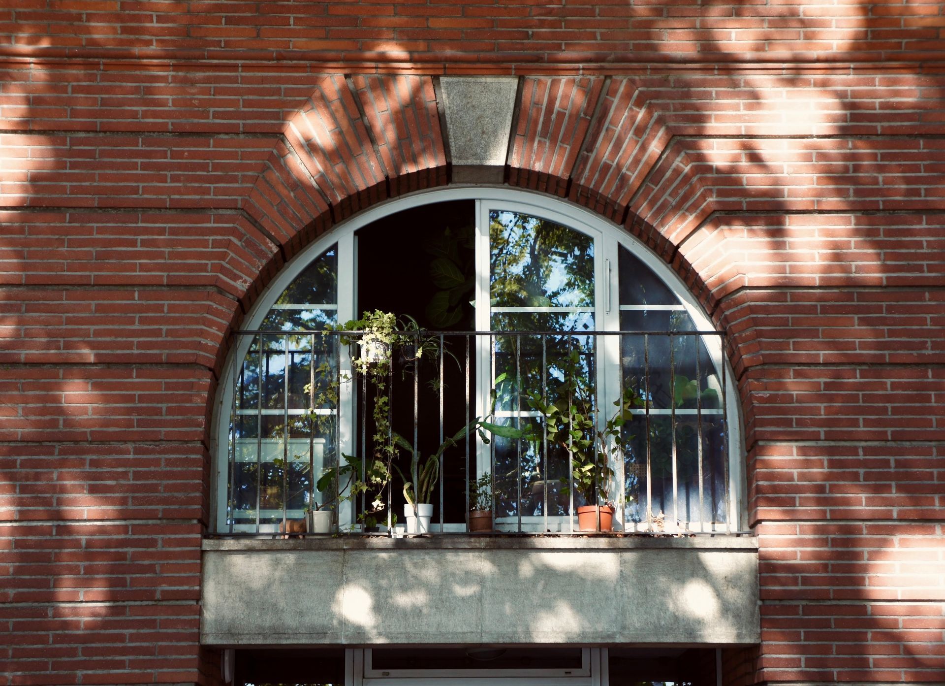 Fenêtre cintrée avec plantes sur un balcon, dans un immeuble en briques rouges ombragé par des arbres.