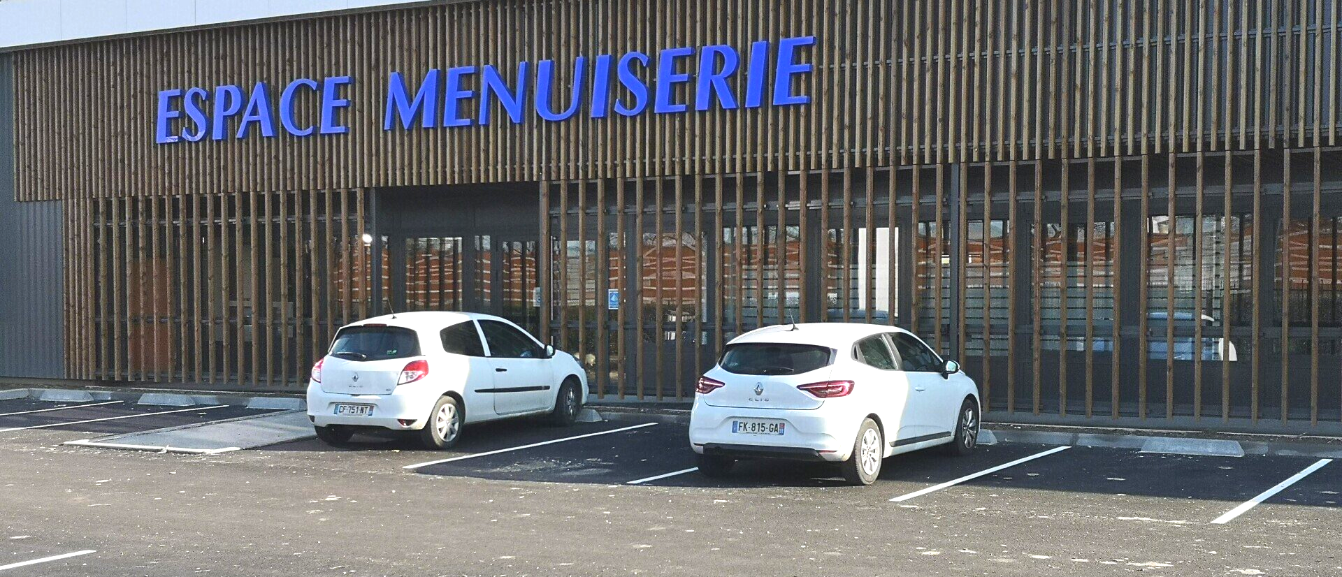 Deux voitures blanches garées devant le bâtiment Espace Menuiserie à façade en bois.
