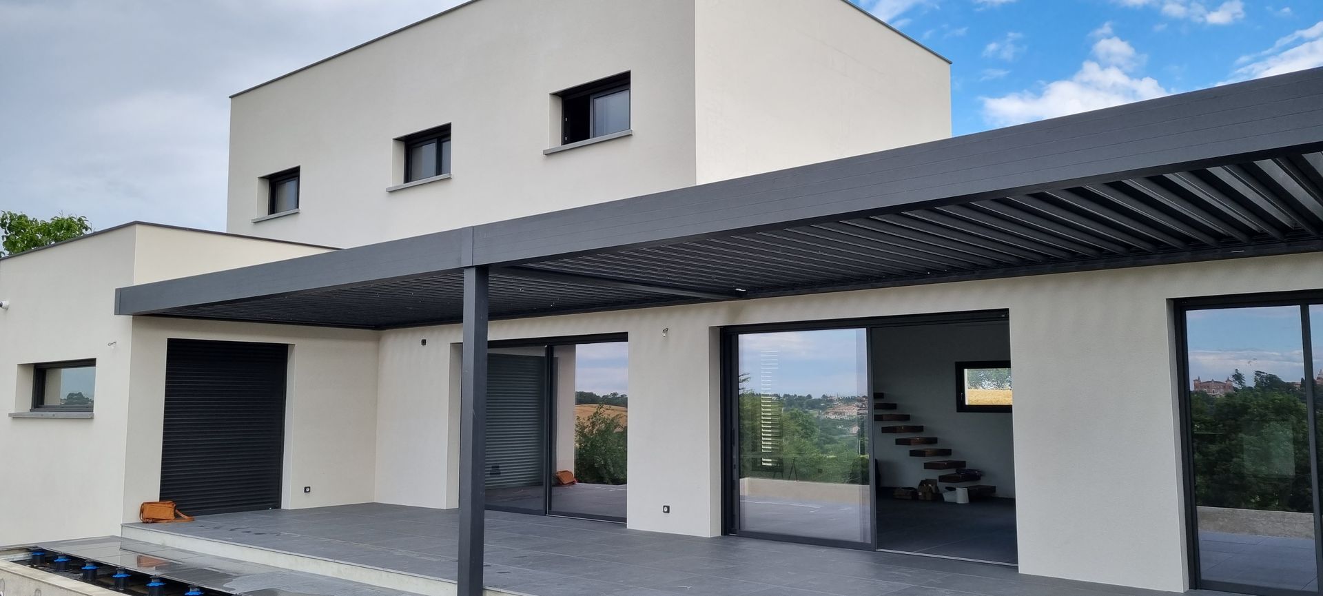 Un abri de voiture en bois attenant à un garage clair avec une porte grise, sous un ciel bleu.