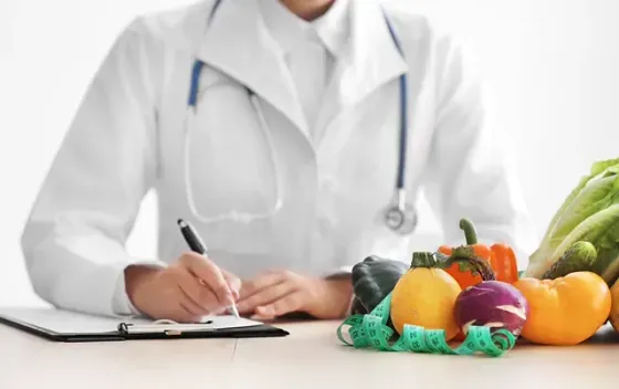 Un médico está sentado en una mesa con frutas y verduras y escribiendo en un portapapeles.