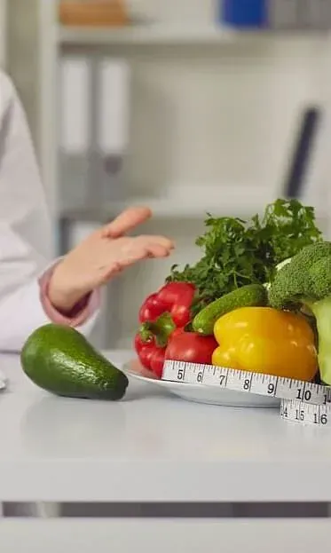 Una mujer está sentada en una mesa con un plato de verduras y una cinta métrica.