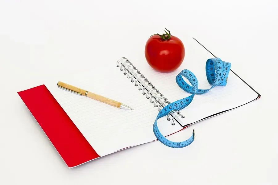 Un tomate, una cinta métrica y un bolígrafo están sobre un cuaderno.