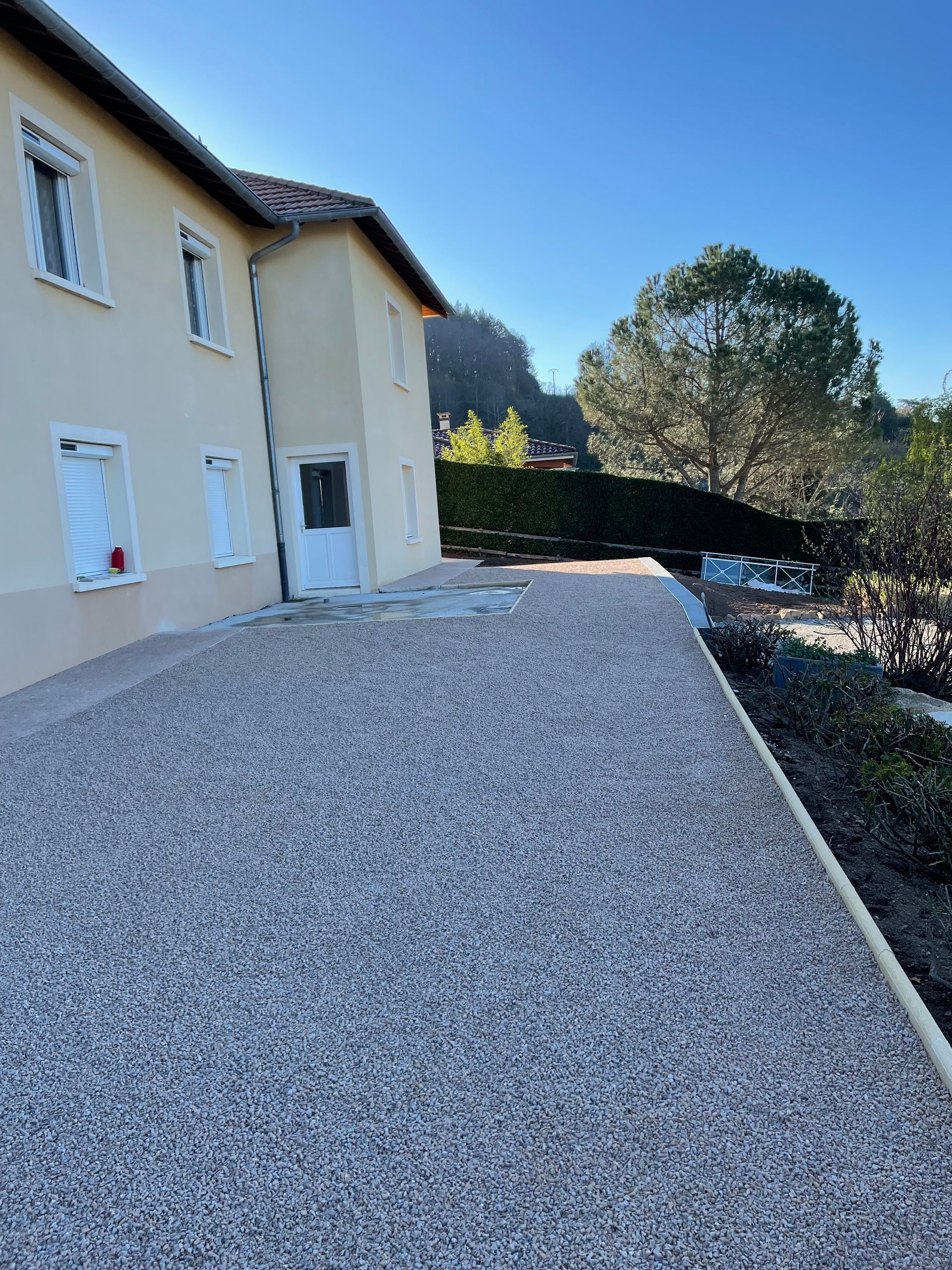 Allée de gravier longeant une maison beige, avec des arbres et des haies sous un ciel bleu azur.
