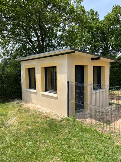 Petit bâtiment annexe beige avec fenêtres sombres et portail métallique, entouré de pelouse et d'arbres.
