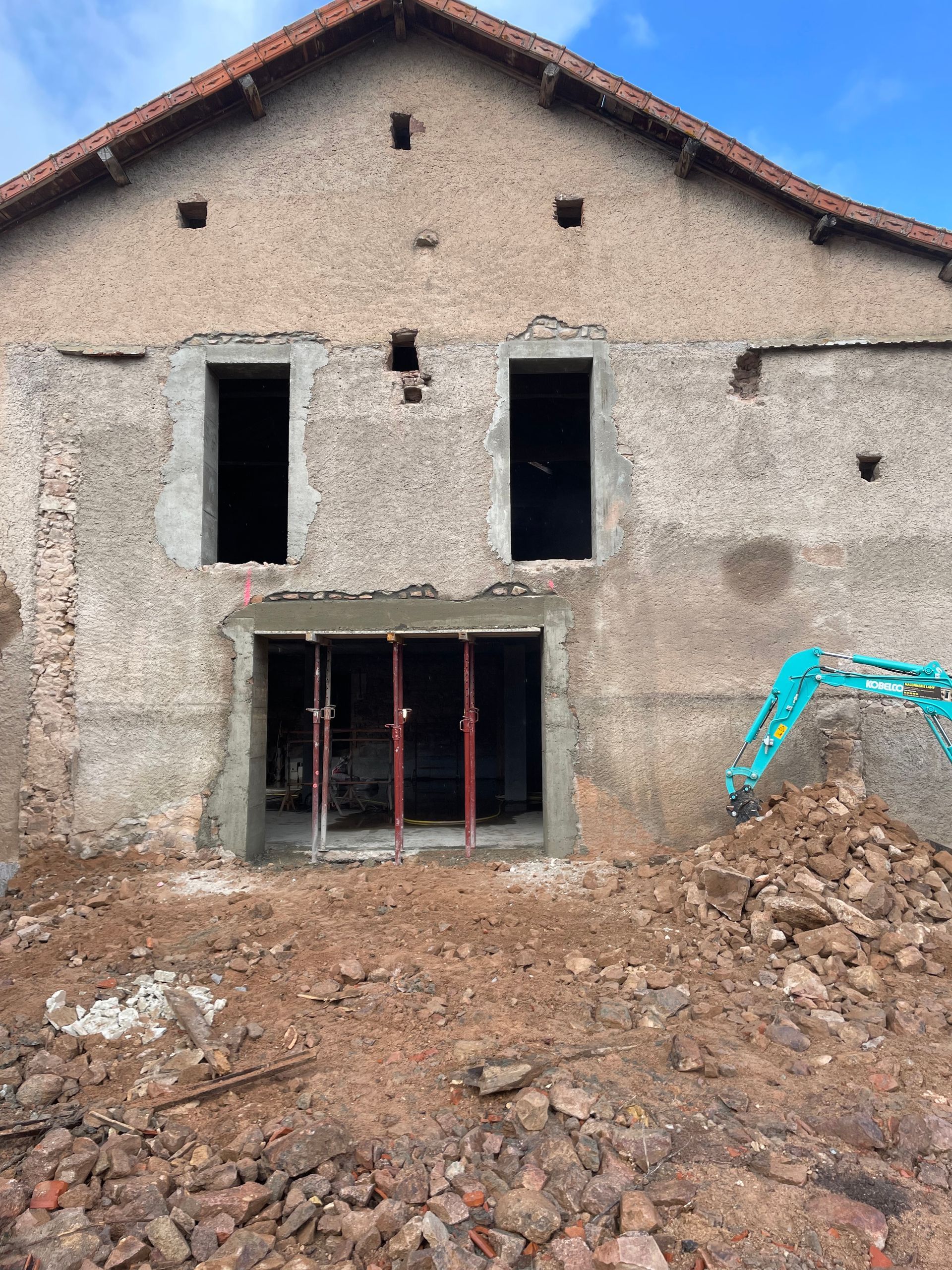 Façade d'un vieux bâtiment en pierre, avec fenêtres et porte ouvertes, entourée de terre et de gravats.