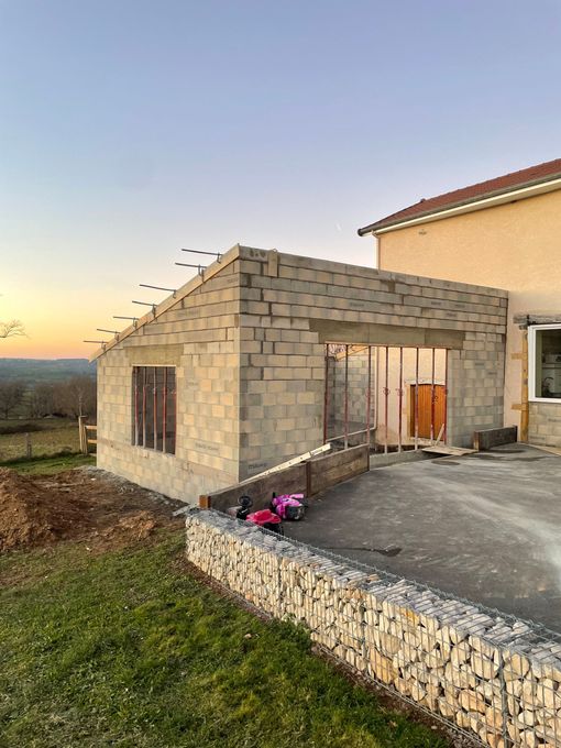 Extension inachevée en parpaings jouxtant une maison beige au coucher du soleil, avec une terrasse en béton et de petites fleurs roses.