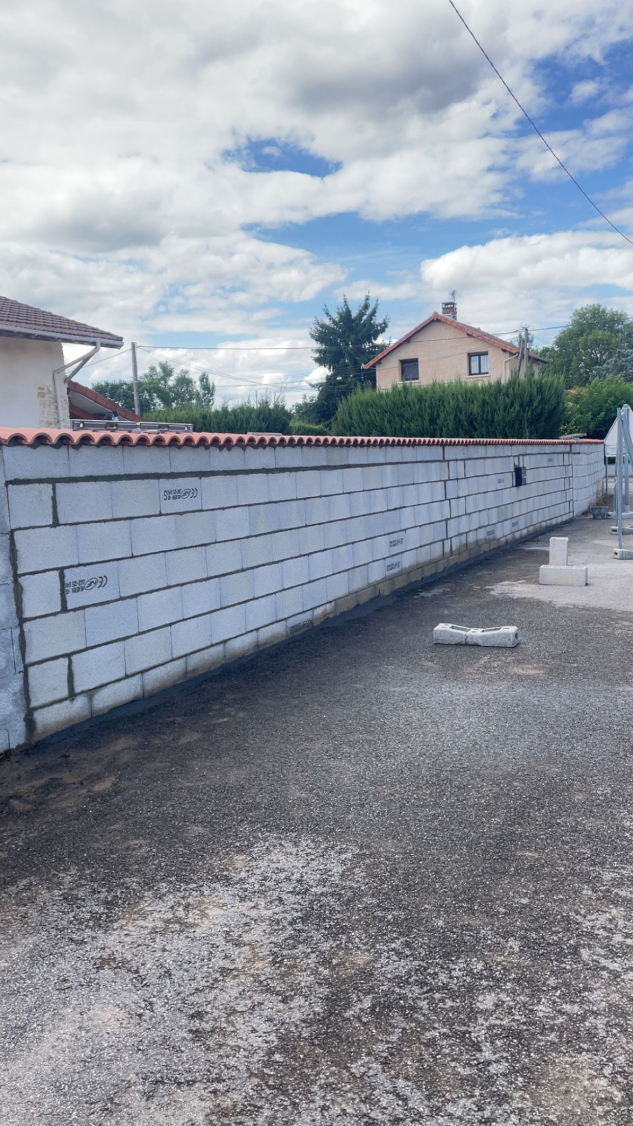 Long mur de maçonnerie blanche bordant une allée de gravier sous un ciel partiellement nuageux dans une cour résidentielle.