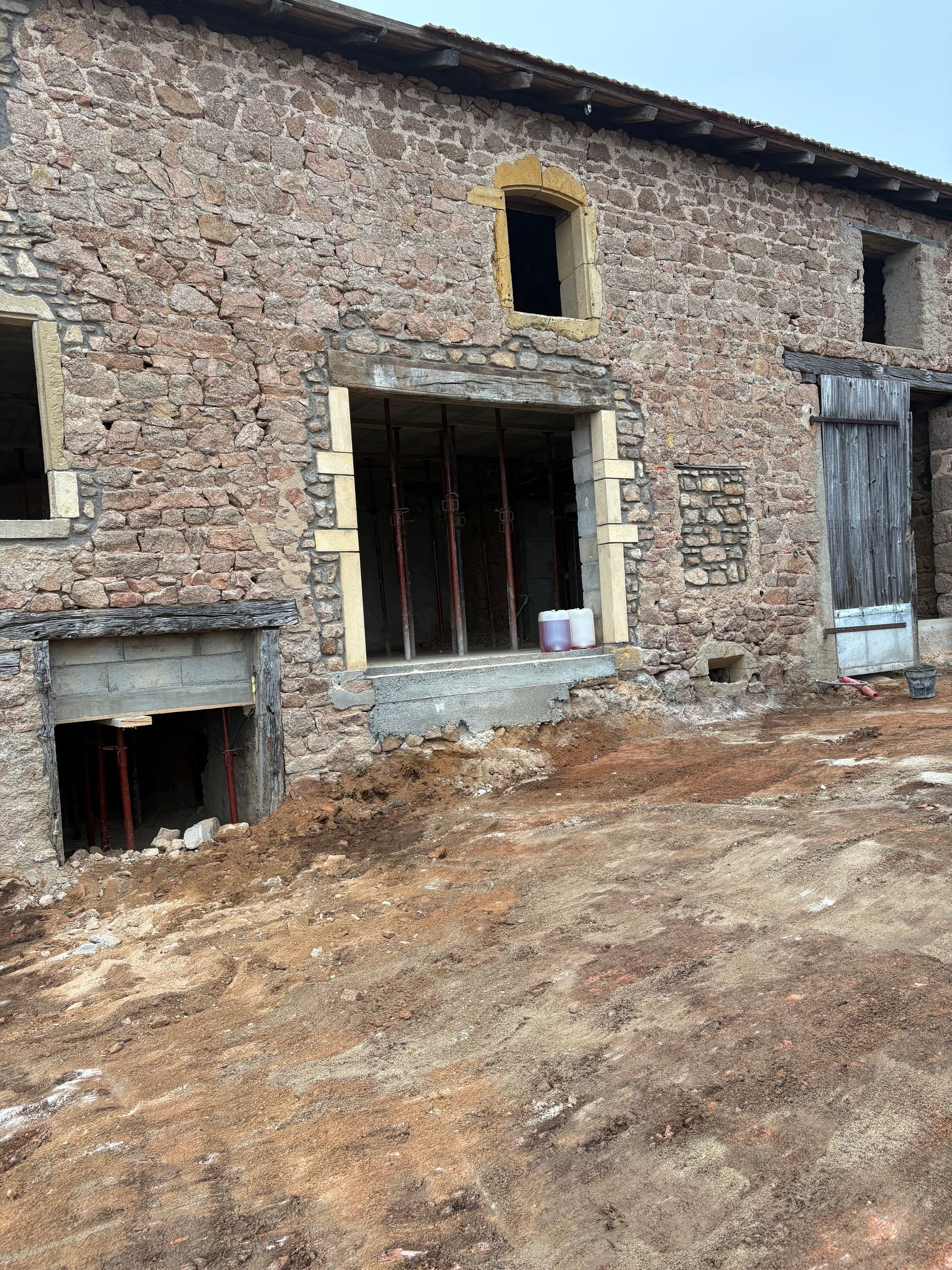 Maison en pierre partiellement construite, avec portes et fenêtres inachevées, sur un terrain en terre battue.
