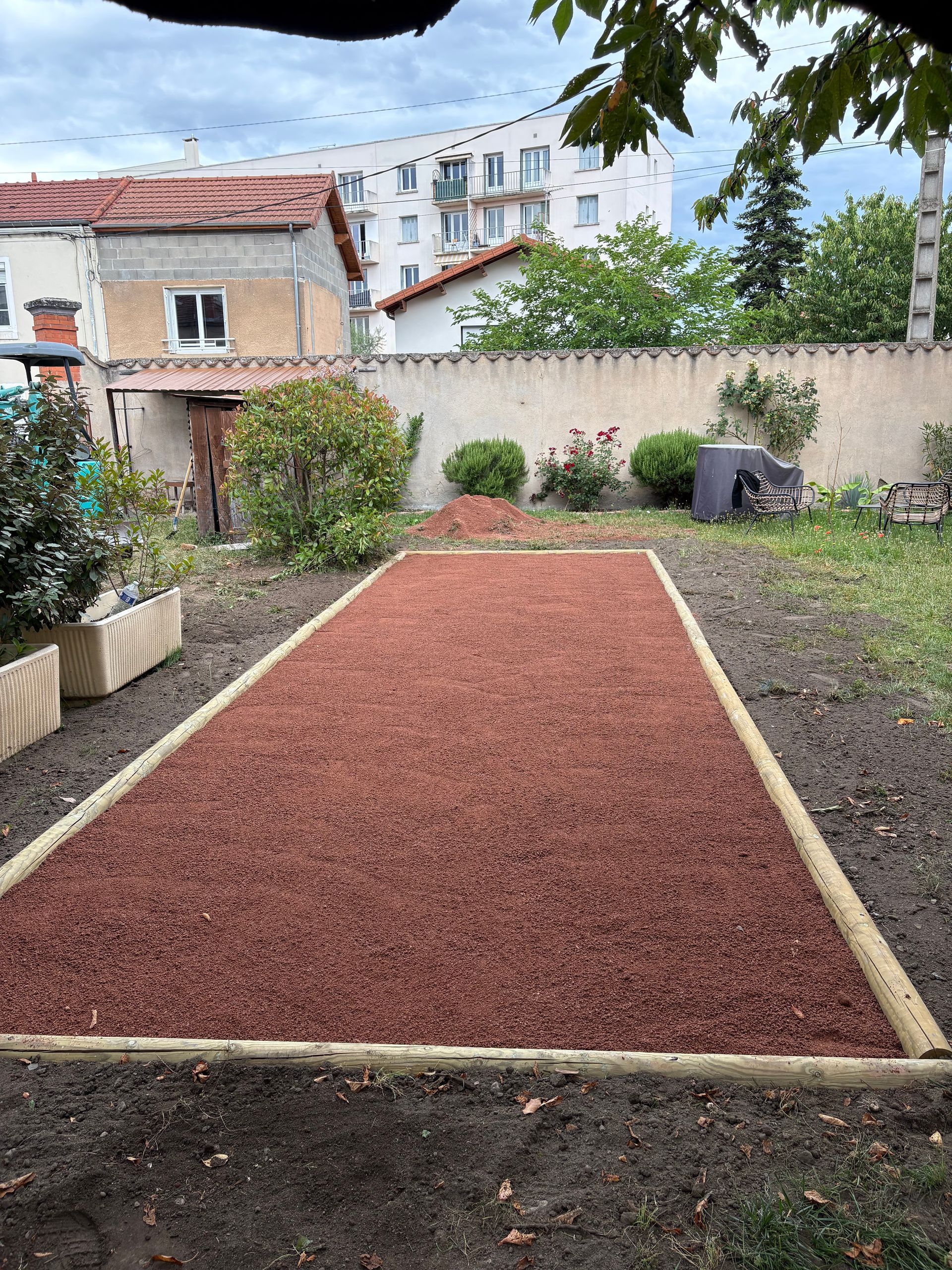 Jardin avec un parterre rectangulaire de gravier rouge bordé de blanc, entouré de plantes et d'un mur.