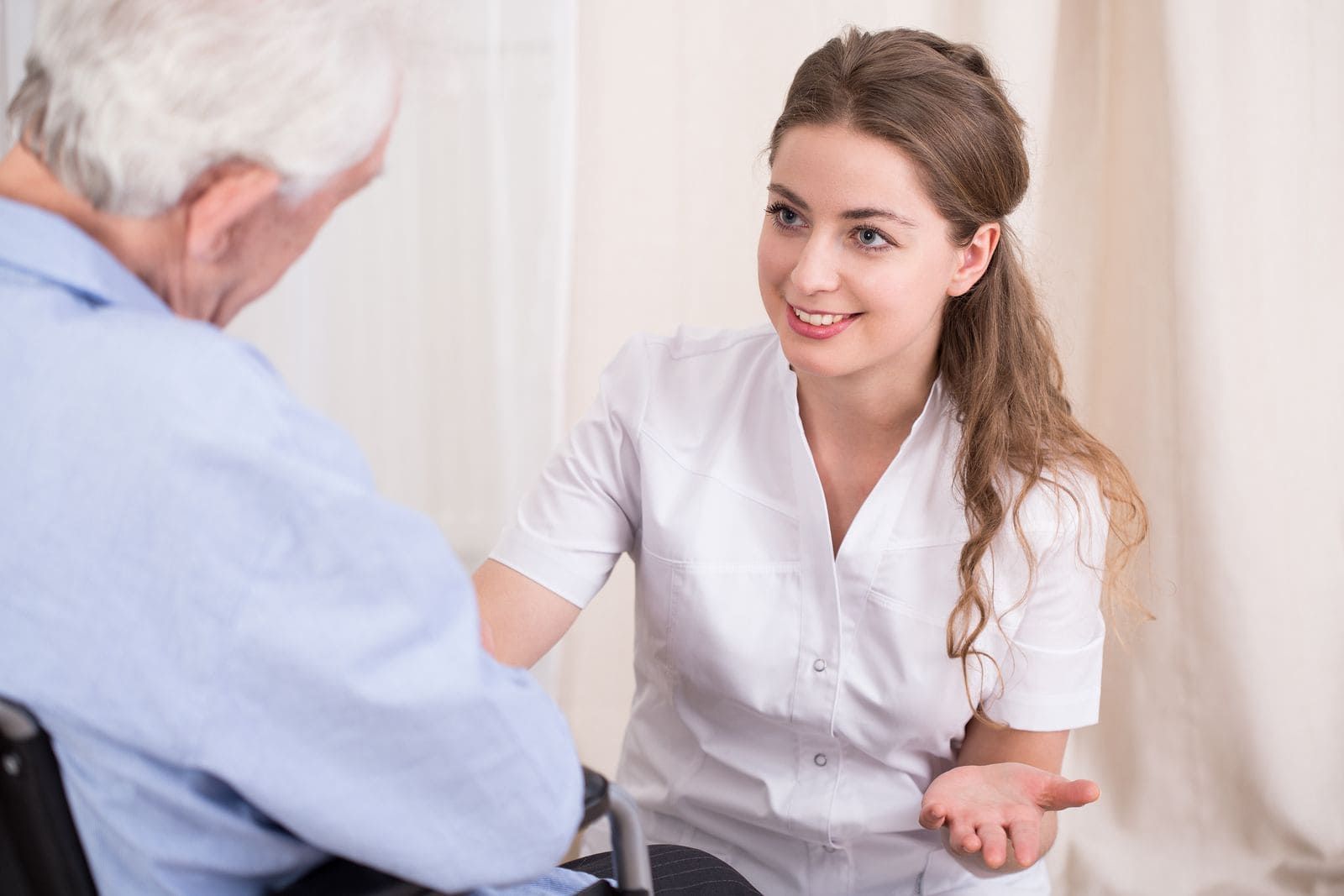 Una mujer con uniforme blanco sonríe mientras habla con una persona en silla de ruedas, en el interior.