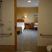 Puerta abierta a un dormitorio. Cama, mesita de noche, escritorio y silla visibles. Paredes y suelo beige.