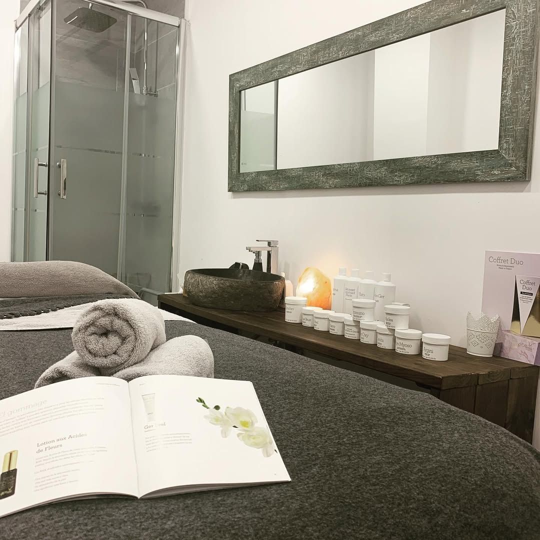 Una sala de tratamiento con una camilla de masaje, toallas grises enrolladas, un libro abierto, un lavabo y productos