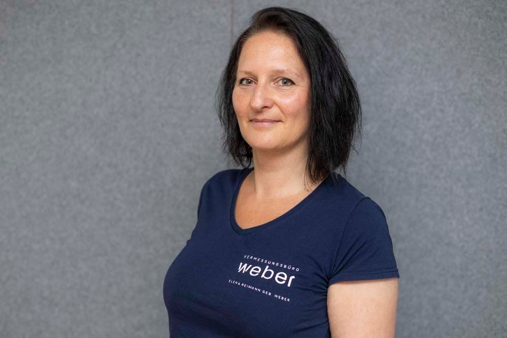 Eine Frau mit dunklem Haar lächelt und trägt ein weißes Hemd