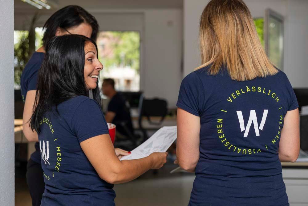 Drei Personen in marineblauen T-Shirts mit einem Logo unterhalten sich in einem Büro. Die Frau vor ihnen lächelt und hält Papiere in der Hand.