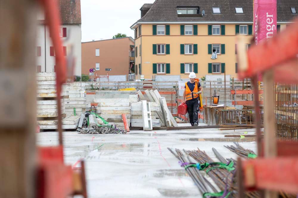 Bauarbeiter in orangefarbener Weste auf einer Baustelle, gelbes Gebäude im Hintergrund.