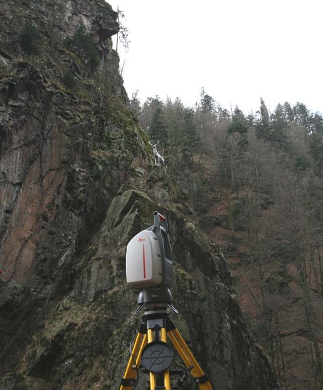 Ein Laserscanner auf einem Stativ untersucht eine felsige Felswand mit spärlichen Bäumen. Bewölkter Himmel.