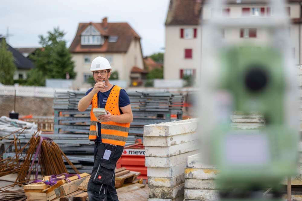 Ein Bauarbeiter mit Schutzhelm und Weste begutachtet eine Baustelle, hält einen Notizblock in der Hand und sieht im Hintergrund Gebäude.