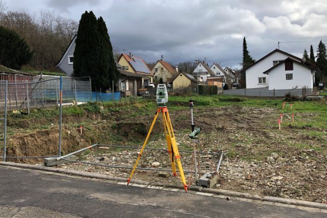 Vermessungsgerät auf einem Stativ auf einer Baustelle, das Land vermessend, mit Häusern im Hintergrund.