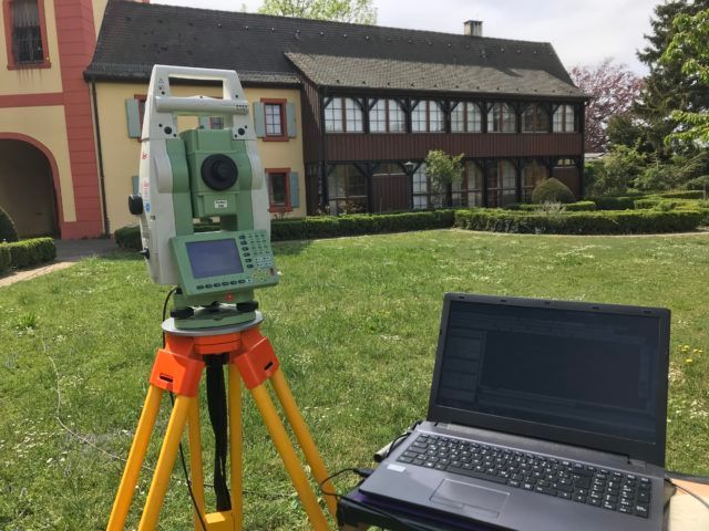 Auf einem Stativ aufgestellte Vermessungsgeräte vor einem mehrstöckigen Haus mit Rasen.