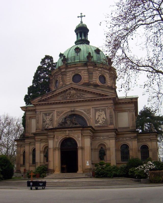 Reich verziertes hellbraunes Mausoleum mit grüner Kuppel und Kreuz vor bewölktem Himmel.