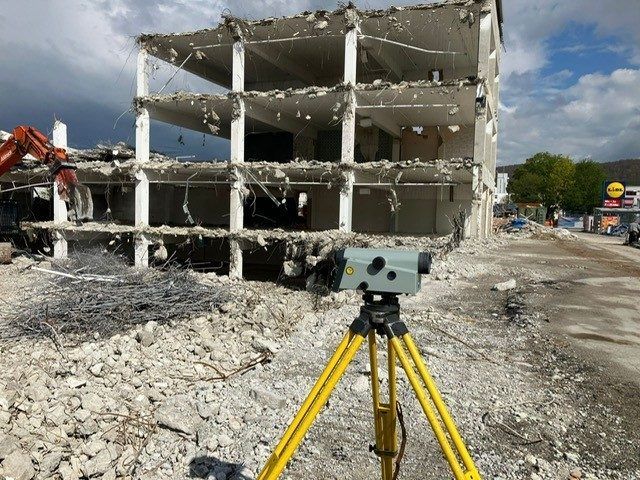 Vermessungsgerät auf einem Stativ vor einem teilweise abgerissenen Gebäude. Schutt liegt verstreut auf dem Boden.