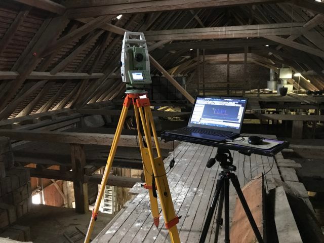 Eine Totalstation zur Vermessung auf einem hölzernen Dachboden. Das Instrument steht auf einem Stativ neben einem Laptop.