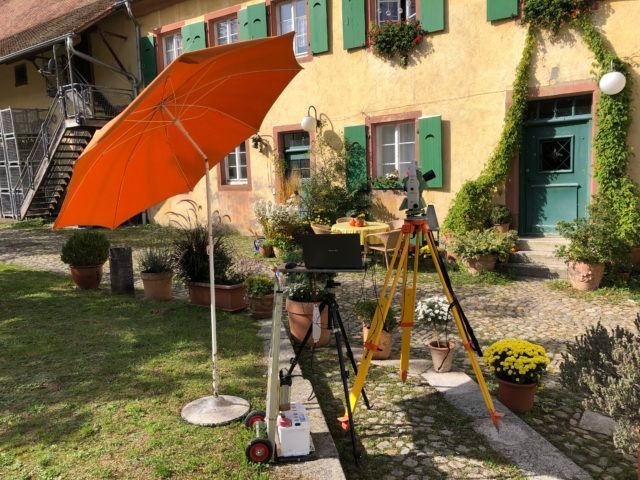 Ein orangefarbener Regenschirm beschattet Vermessungsgeräte vor einem gelben Gebäude mit grünen Fensterläden und einer grünen Tür.