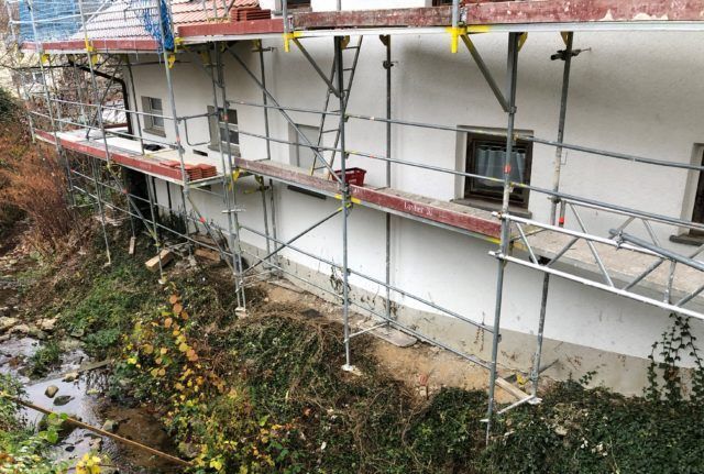 Gebäude mit Gerüst; Außenrenovierung im Gange; darunter überwucherte Vegetation.