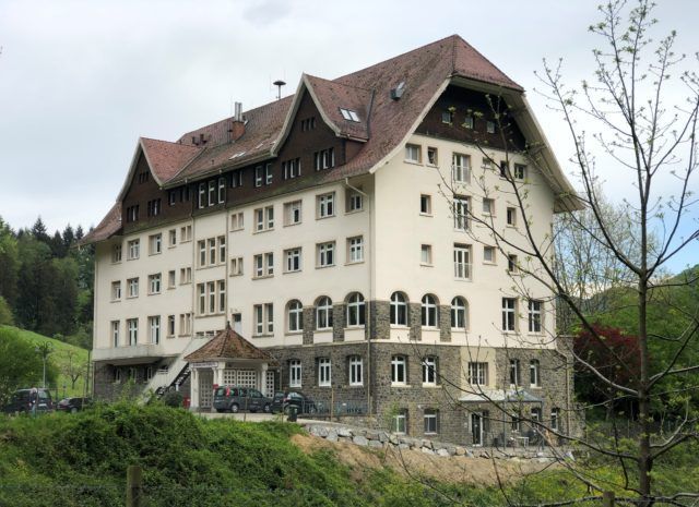 Großes, weißes Gebäude mit rotem Satteldach und Steinsockel. In der Nähe des Eingangs parken Autos, umgeben von einer ländlichen Landschaft.
