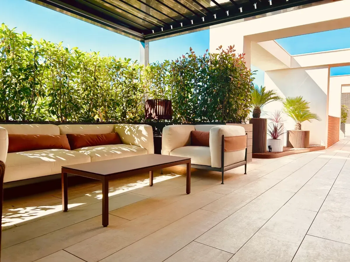 Patio moderno con sofá, mesa de centro, sillas y plantas en macetas bajo una pérgola sombreada.