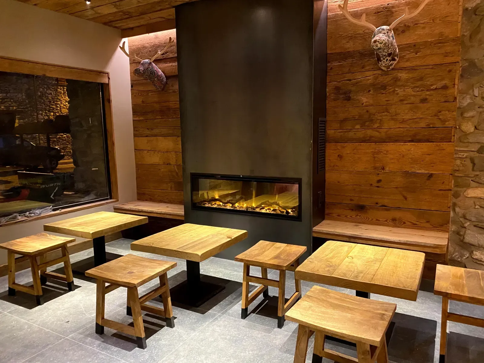 Acogedor salón con chimenea de piedra, paredes de madera y mesas bajas y taburetes distribuidos por toda la estancia.