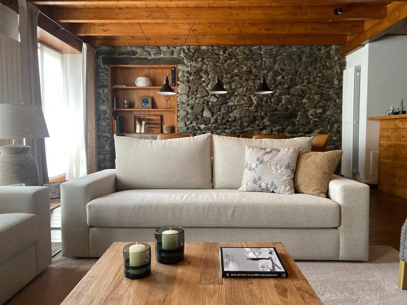 Acogedora sala de estar con sofá beige, techo de madera, pared decorativa de piedra y velas en la mesa de centro.
