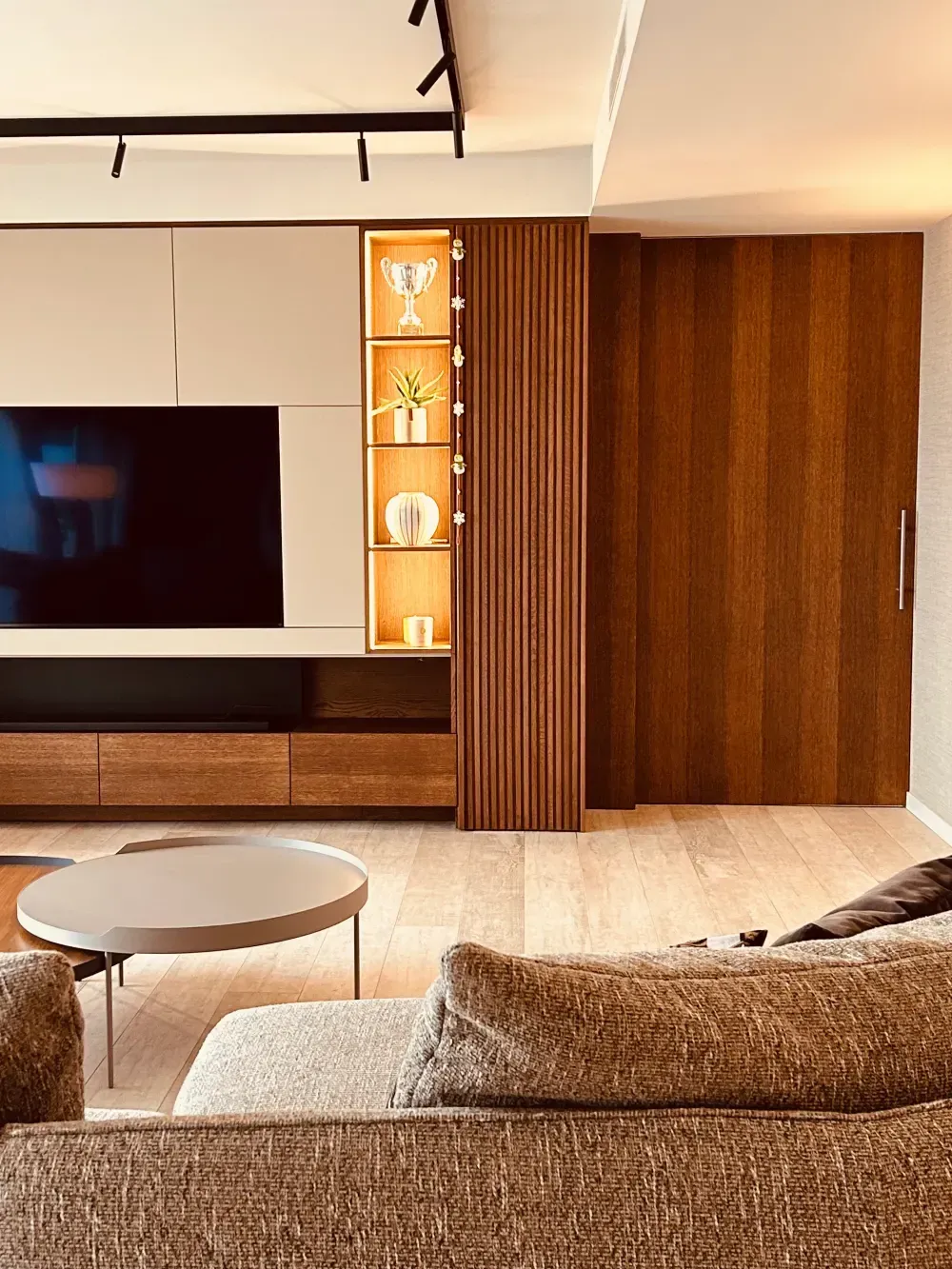 Sala de estar moderna con televisor montado en la pared, armarios de madera, sofá beige, mesa redonda e iluminación cálida.