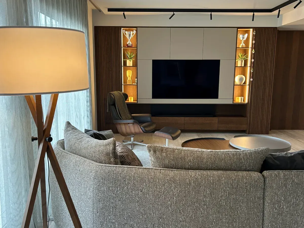 Sala de estar moderna con sofá gris, televisor grande, lámpara de pie y estantes decorativos.
