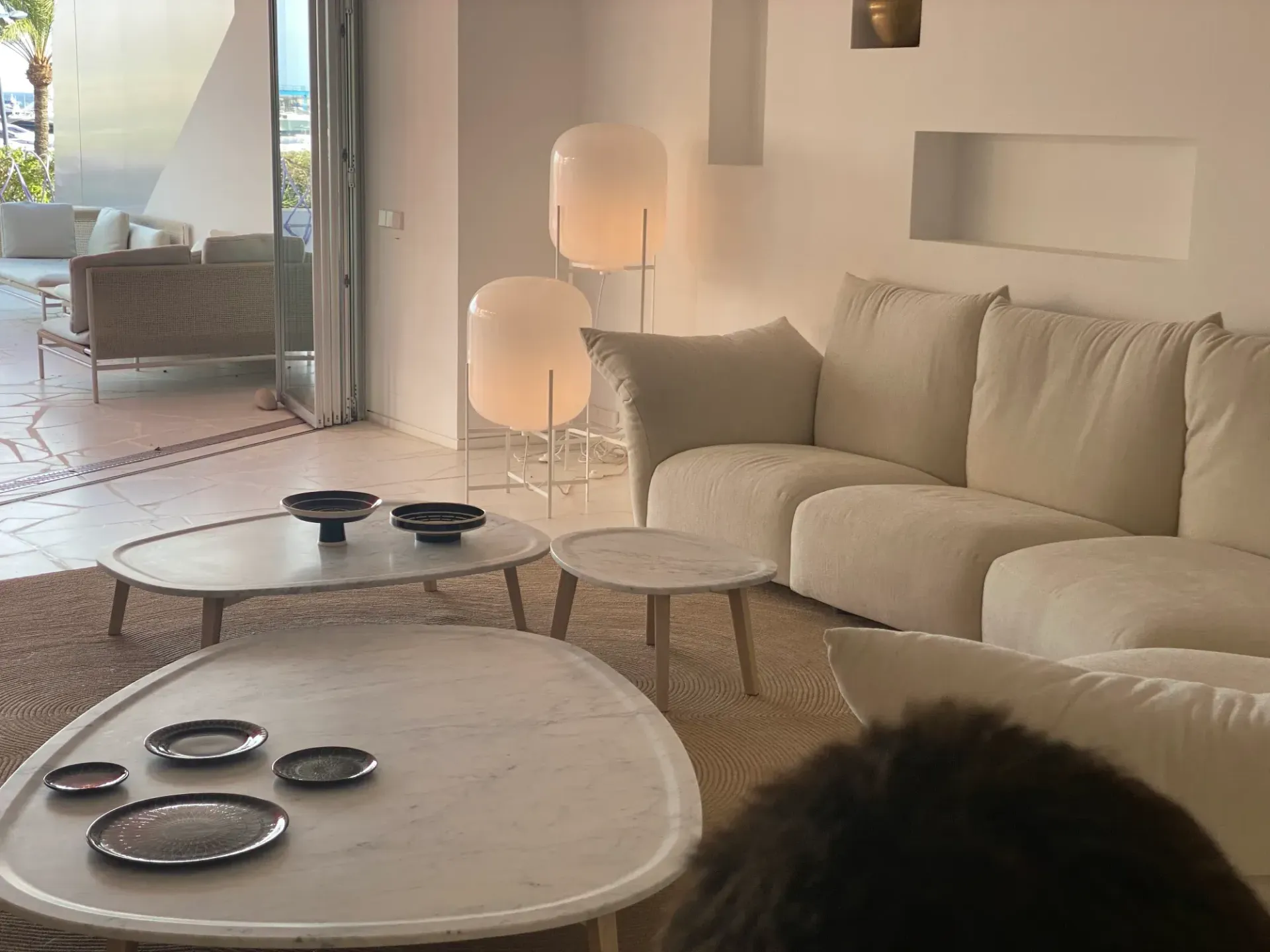 Acogedora sala de estar con sofá beige, mesas redondas y lámparas cálidas, con acceso a un luminoso patio.