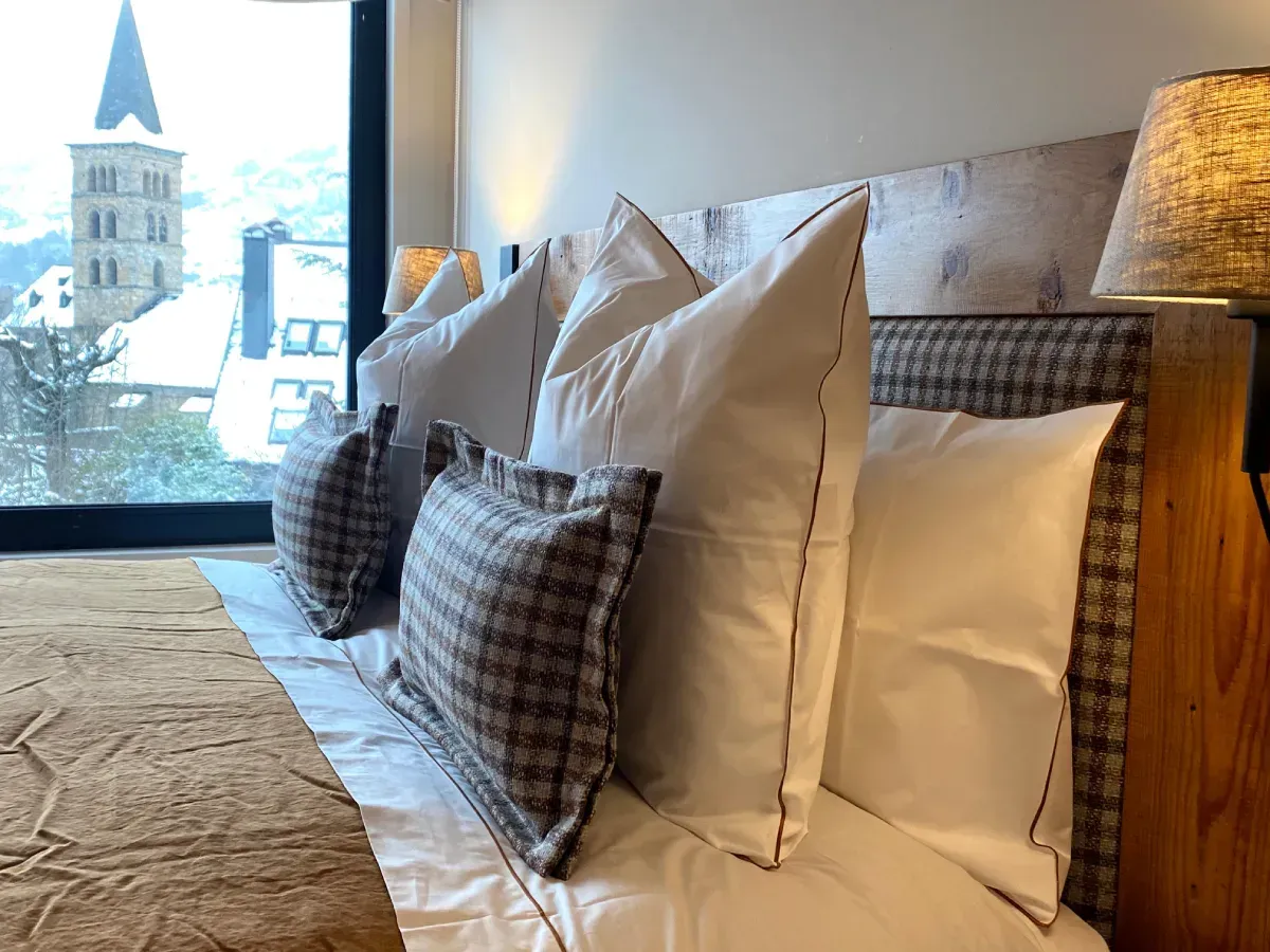 Cama de hotel bien hecha con almohadas blancas y a cuadros junto a una ventana y una lámpara de noche.