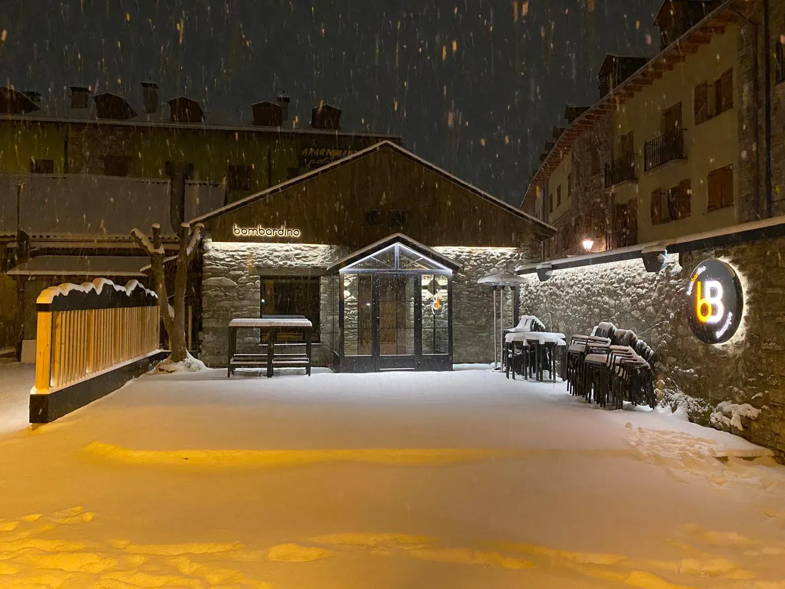 Patio nevado por la noche con un cobertizo iluminado, un muro de piedra y un letrero luminoso bajo la nieve que cae.
