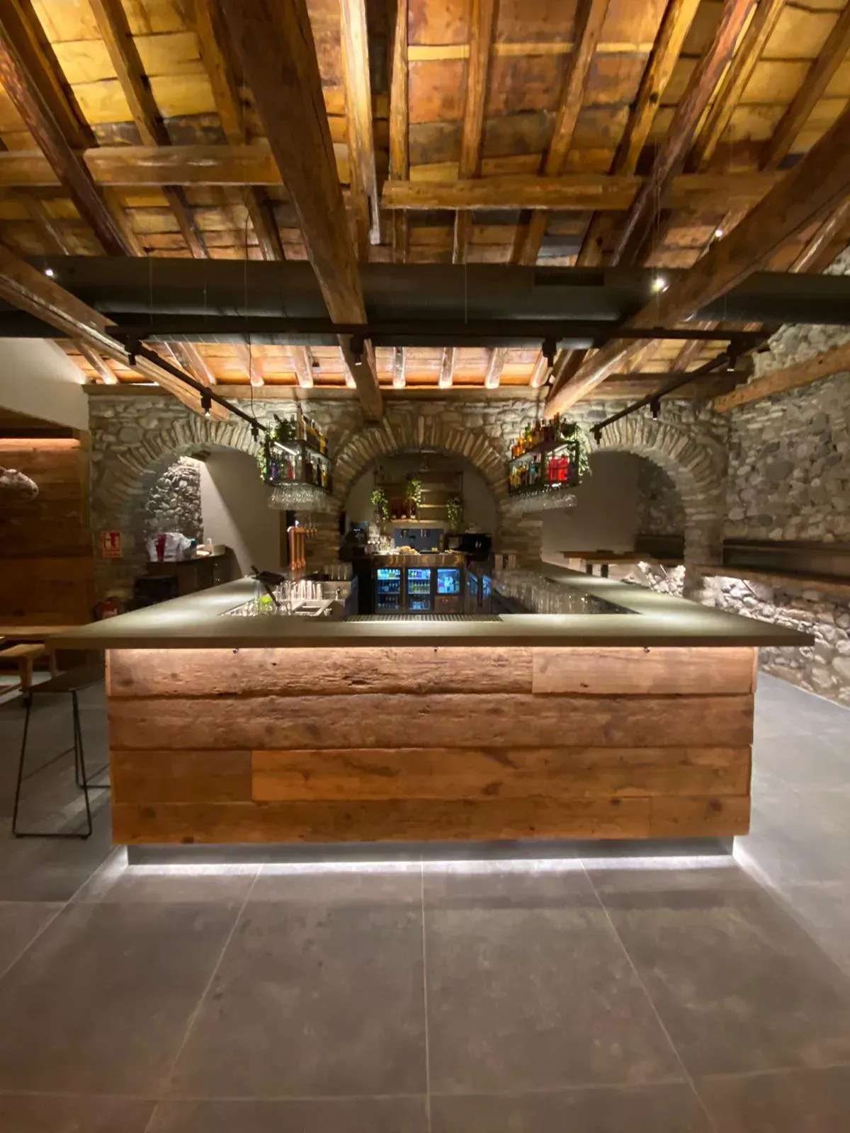 Interior de bar rústico con vigas de madera, arcos de piedra y una barra central iluminada desde abajo.