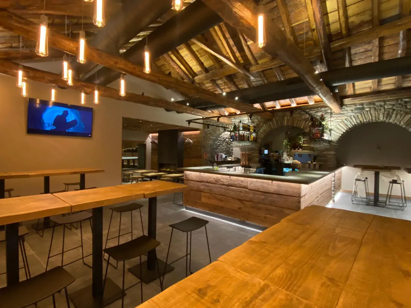 Interior de bar rústico vacío con vigas de madera, taburetes y un televisor en la pared.