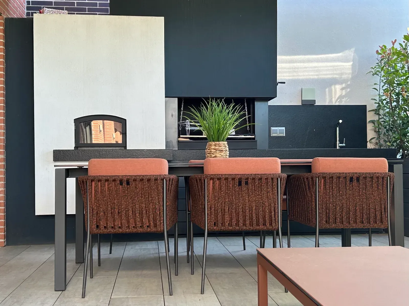 Patio exterior moderno con taburetes de bar, mostrador negro, chimenea y planta en maceta sobre una mesa.