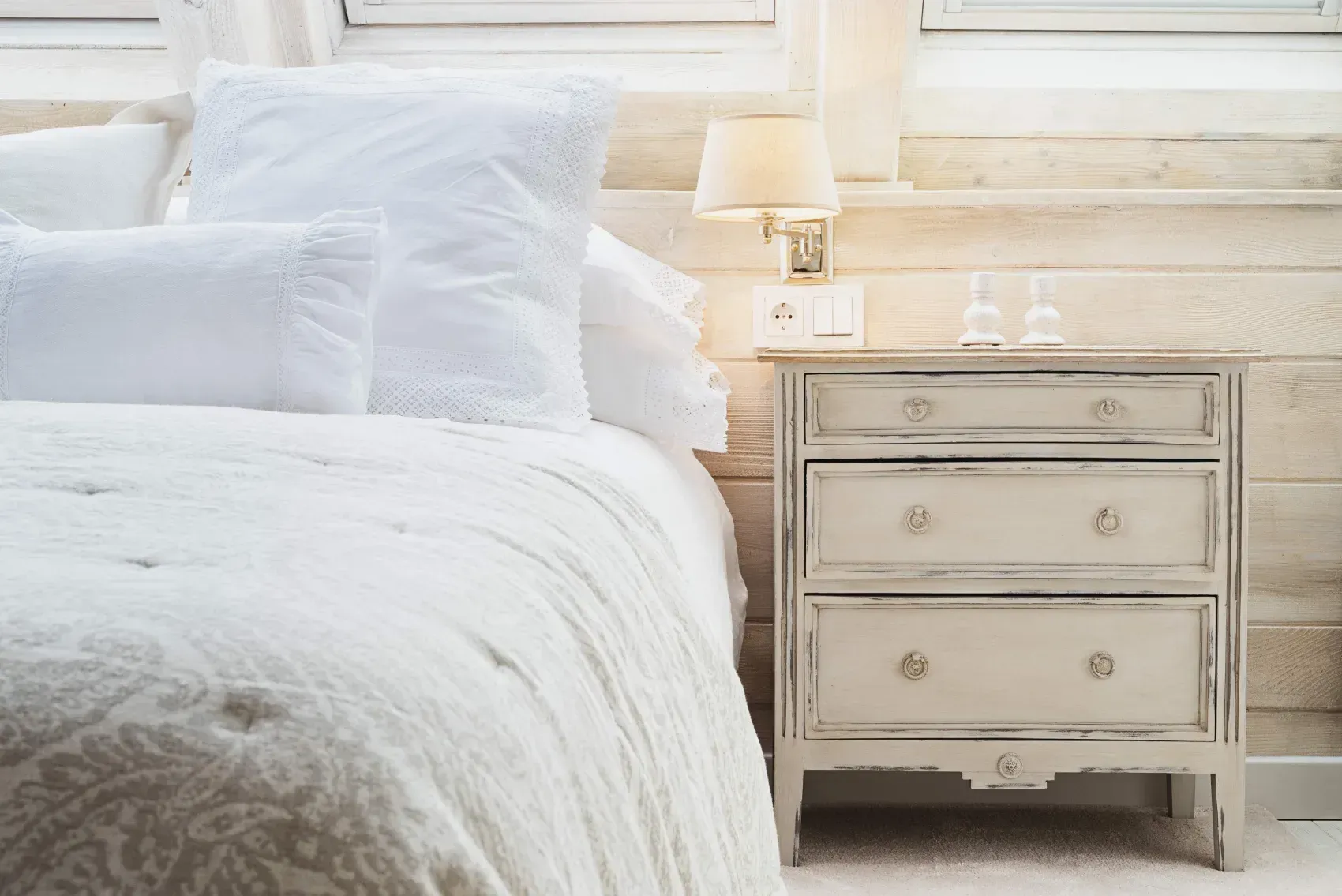 Cama blanca con almohadas junto a una mesita de noche color crema y una lámpara en un dormitorio luminoso.