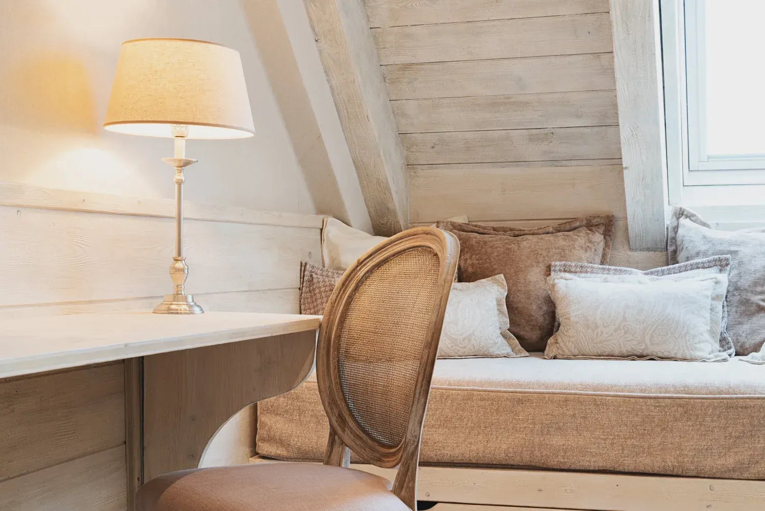 Acogedor rincón de lectura en tonos beige con escritorio, lámpara, silla de mimbre y asiento acolchado junto a la ventana.